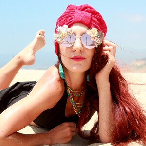 May include: A woman with red hair wearing a red turban, black swimsuit, and large sunglasses with floral embellishments. She is lying on a sandy beach.