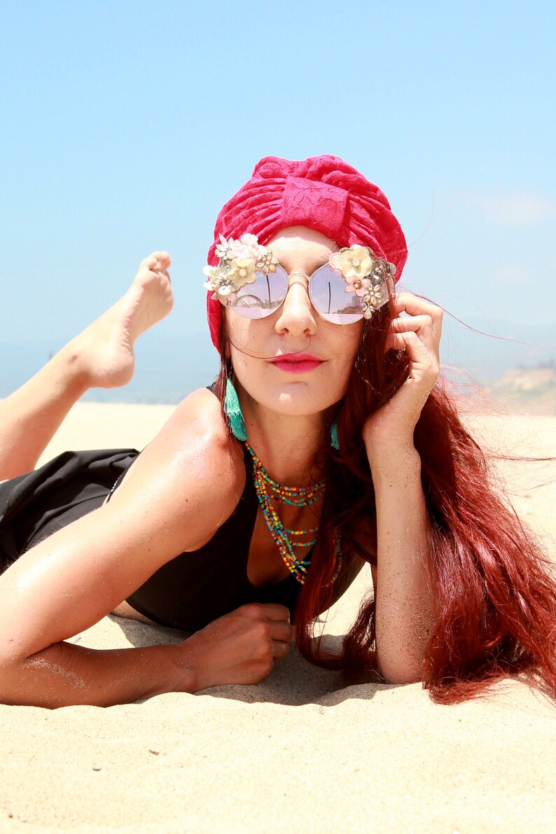 May include: A woman with red hair wearing a red turban, black swimsuit, and large sunglasses with floral embellishments. She is lying on a sandy beach.