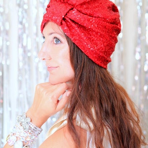 Puede incluir: Una mujer con el pelo largo, ondulado y casta&ntilde;o rojizo lleva un turbante rojo con lentejuelas. Lleva una blusa blanca y varias pulseras en la mu&ntilde;eca.