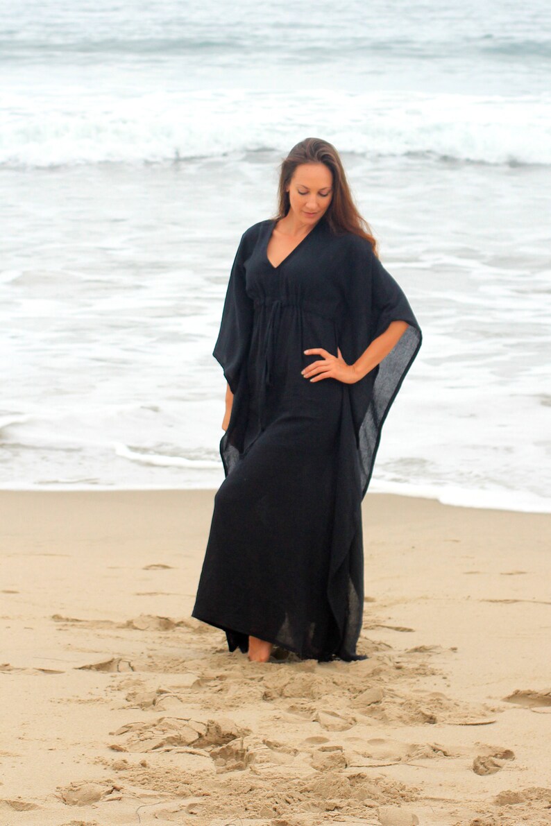 Puede incluir: Una mujer con un vestido largo de caft&aacute;n negro y fluido en una playa de arena. El vestido tiene un escote en V y una cintura con cord&oacute;n.
