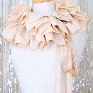 May include: A large, cream-coloured, ruffled collar with a matching satin ribbon tie. The collar is displayed on a white mannequin.