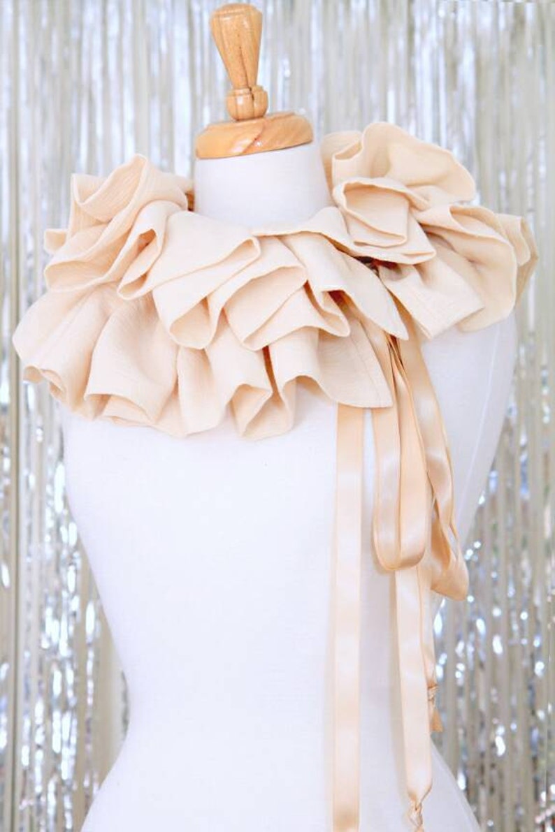 May include: A large, cream-coloured, ruffled collar with a matching satin ribbon tie. The collar is displayed on a white mannequin.