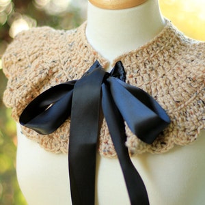 May include: A crocheted collar with a ruffled edge in a beige and brown color with a black satin ribbon tied in a bow.
