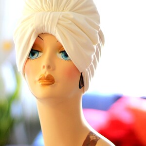 May include: A white turban style head wrap with a large bow on top. The head wrap is made of a soft, smooth fabric. The head wrap is on a mannequin with a painted face and a tattoo on the shoulder.