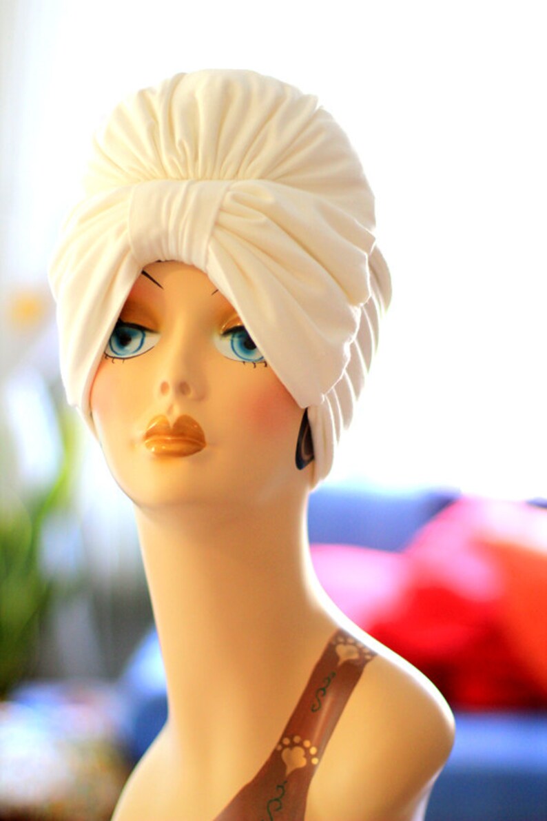 May include: A white turban style head wrap with a large bow on top. The head wrap is made of a soft, smooth fabric. The head wrap is on a mannequin with a painted face and a tattoo on the shoulder.