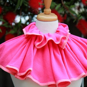 May include: A bright pink ruffled collar with a light pink trim. The collar is displayed on a white mannequin.