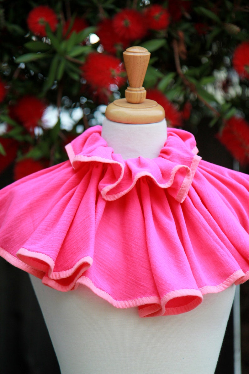 May include: A bright pink ruffled collar with a light pink trim. The collar is displayed on a white mannequin.