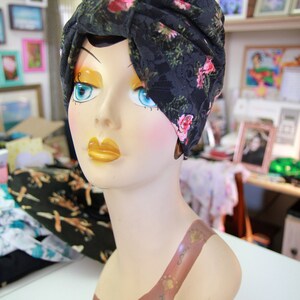 Puede incluir: Un turbante de encaje negro con estampado floral rosa. El turbante est&aacute; en un maniqu&iacute; con un dise&ntilde;o pintado marr&oacute;n en el pecho.