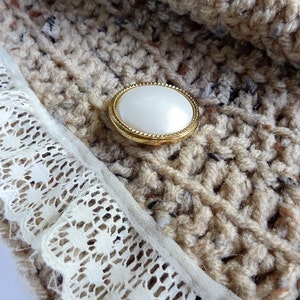 May include: Close-up of a beige crocheted fabric with a white lace trim and two pearl-like buttons with gold rims.