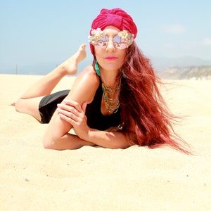 May include: A woman with long red hair is lying on a sandy beach. She is wearing a red turban, mirrored sunglasses with floral embellishments, a black tank top, and a beaded necklace.
