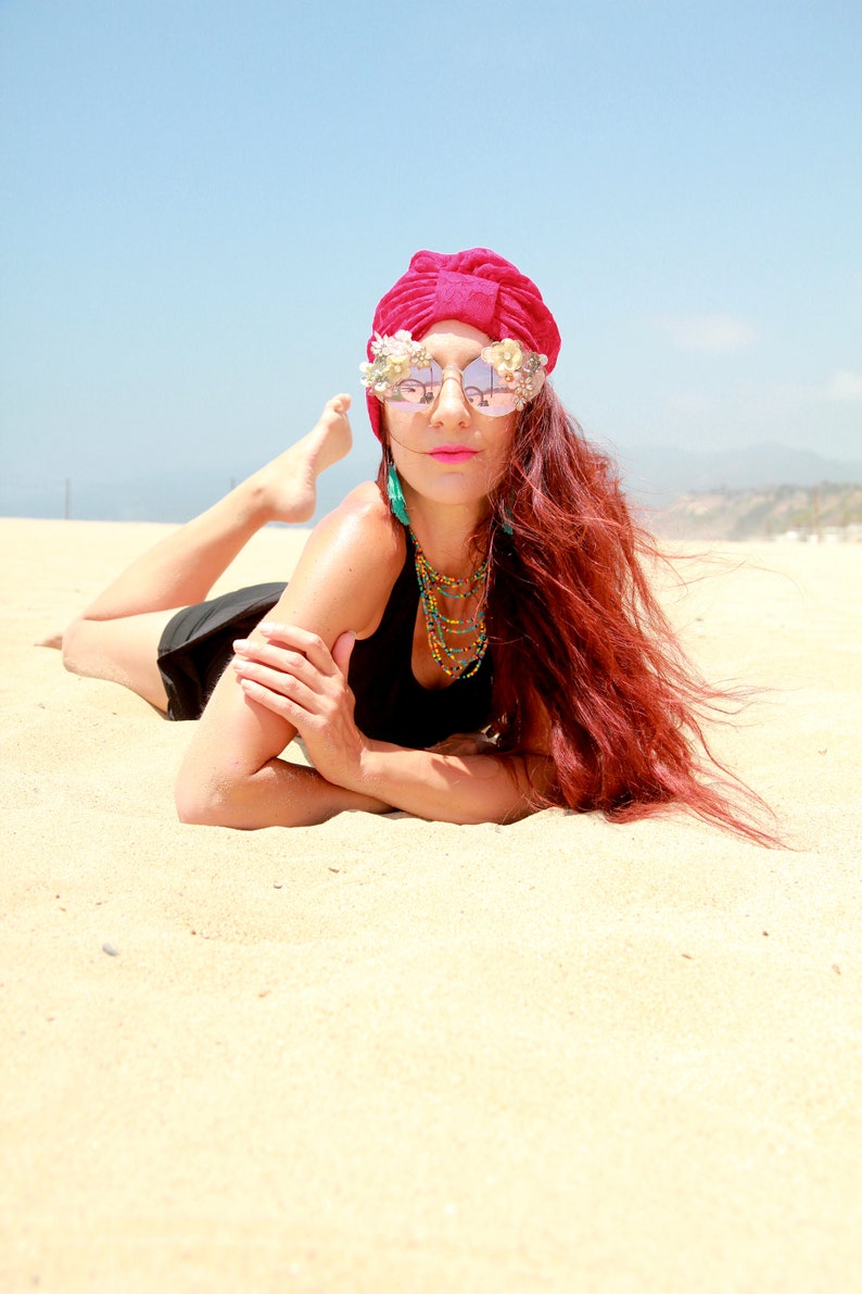 May include: A woman with long red hair is lying on a sandy beach. She is wearing a red turban, mirrored sunglasses with floral embellishments, a black tank top, and a beaded necklace.
