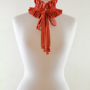 May include: A terracotta-colored ruffled collar with a matching ribbon tie. The collar is displayed on a white mannequin.