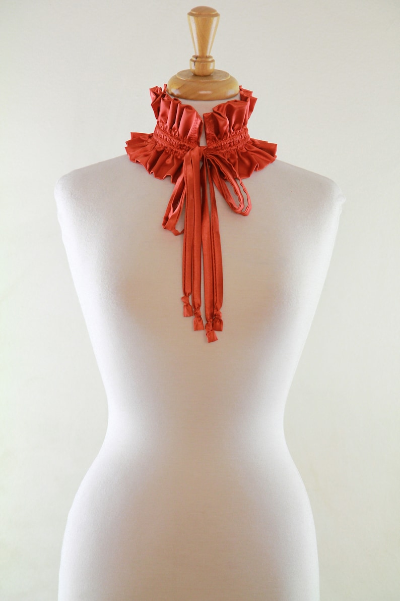 May include: A terracotta-colored ruffled collar with a matching ribbon tie. The collar is displayed on a white mannequin.
