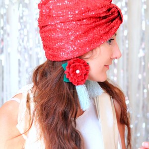 Puede incluir: Una mujer con un turbante rojo de lentejuelas con una gran flor roja y un pendiente de borla azul. Lleva una blusa blanca y tiene el pelo largo y casta&ntilde;o.
