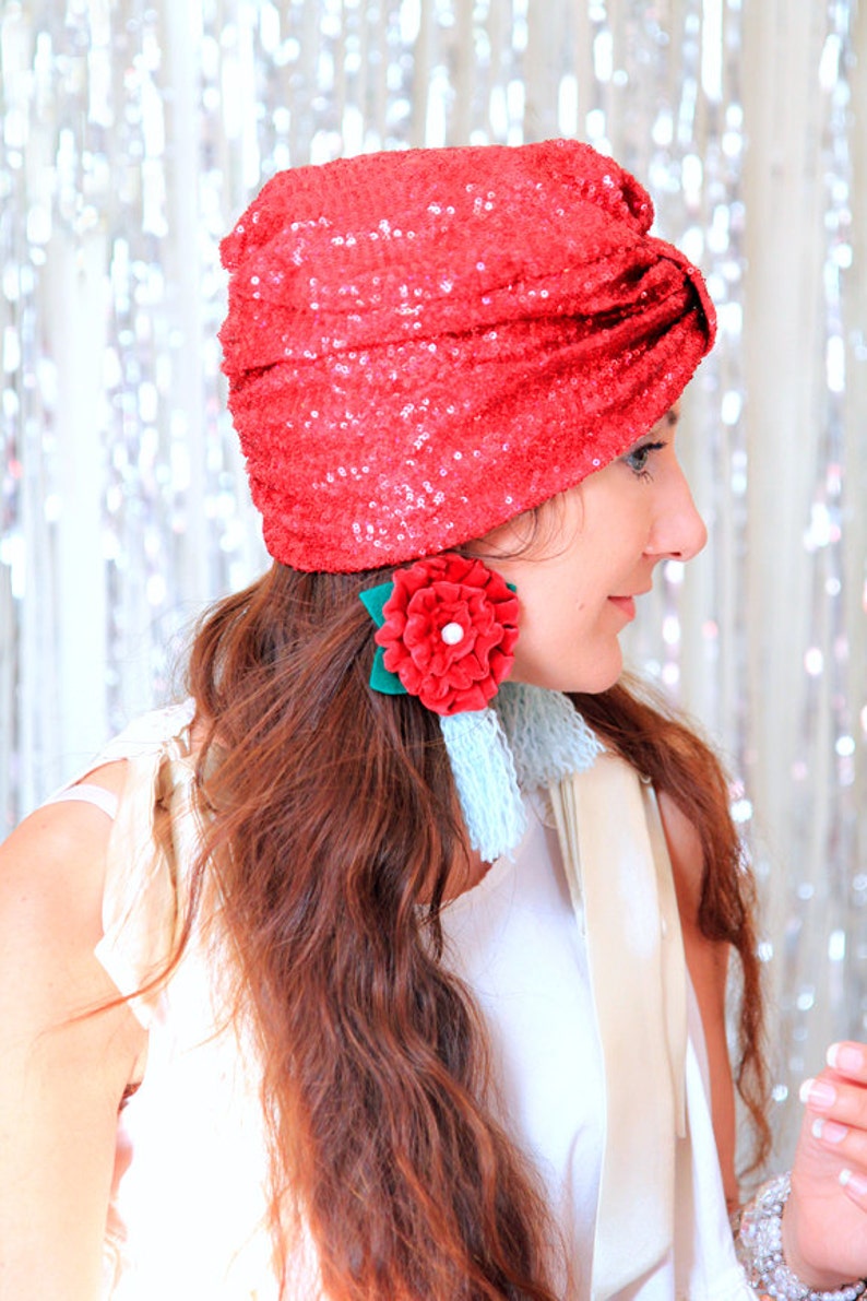 Puede incluir: Una mujer con un turbante rojo de lentejuelas con una gran flor roja y un pendiente de borla azul. Lleva una blusa blanca y tiene el pelo largo y casta&ntilde;o.