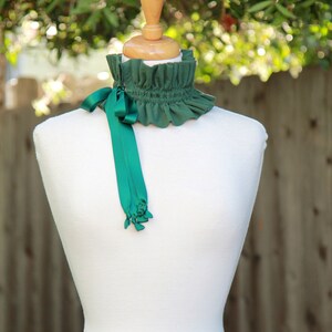 May include: A green ruffled collar with a matching green satin ribbon tied in a bow. The collar is displayed on a white mannequin.