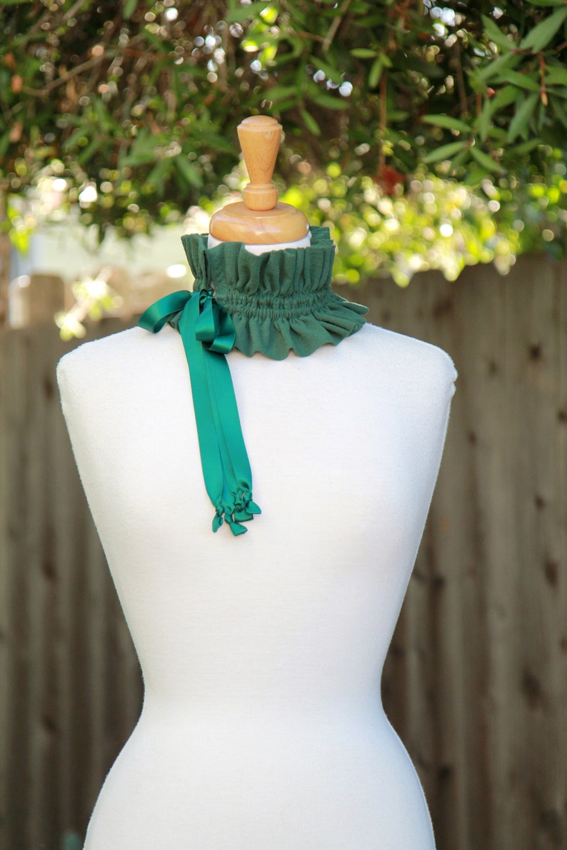 May include: A green ruffled collar with a matching green satin ribbon tied in a bow. The collar is displayed on a white mannequin.