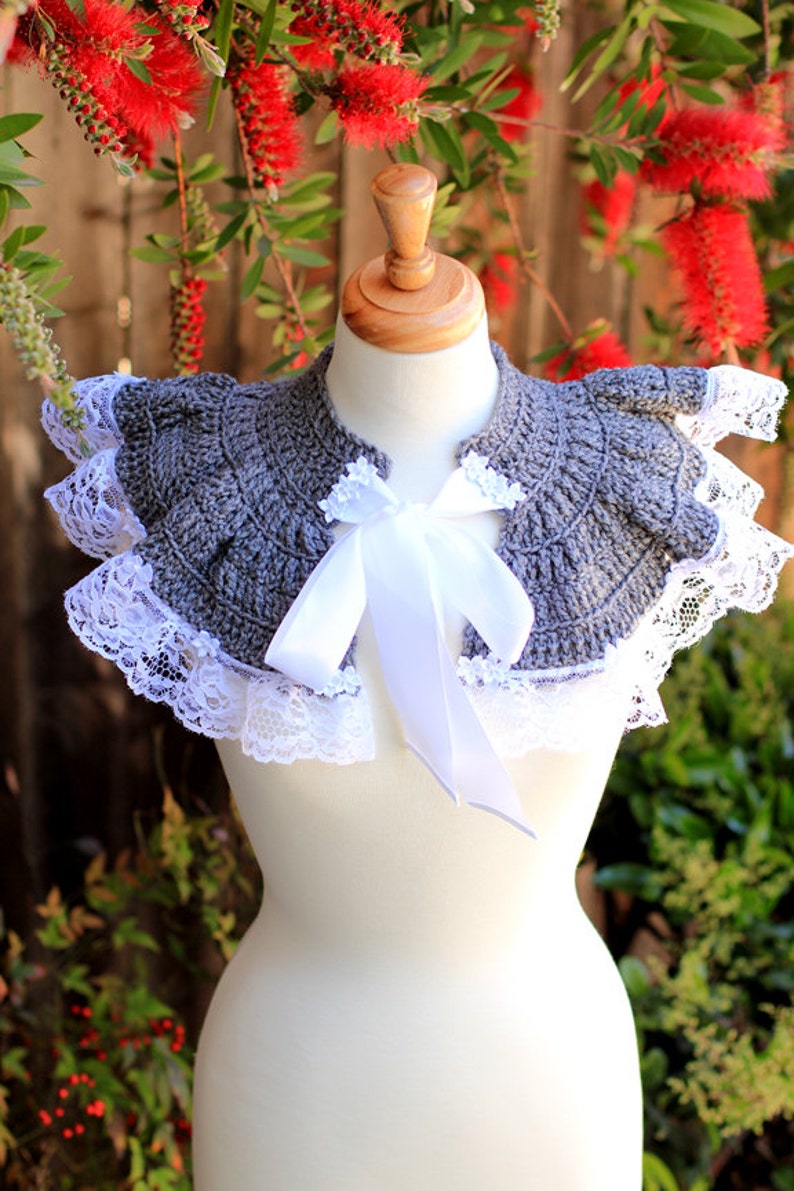 May include: A gray crocheted collar with white lace trim and a white satin ribbon tied in a bow. The collar is displayed on a white mannequin.