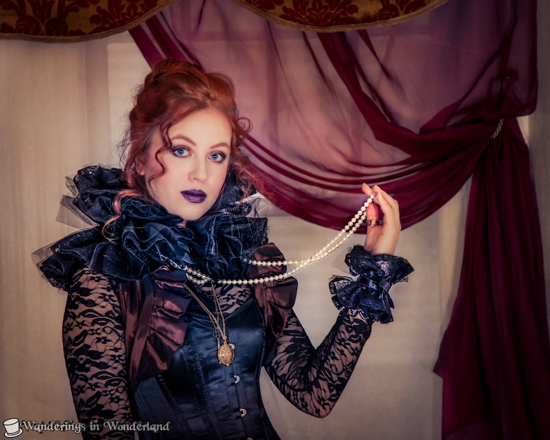 May include: A woman with red hair wearing a black lace top with a large ruffled collar and a pearl necklace. She is standing in front of a red and gold curtain.