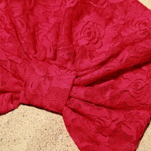 May include: A red lace turban with a floral pattern. The turban is tied in a knot at the top and has a bow on the side.