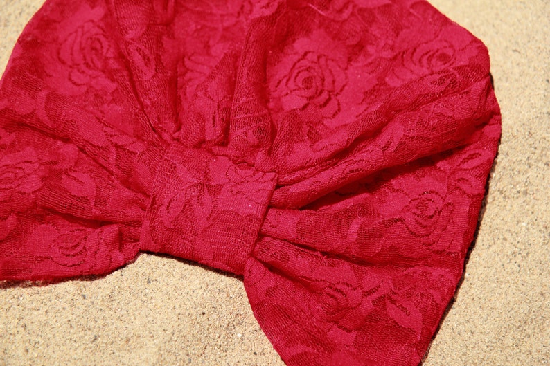 May include: A red lace turban with a floral pattern. The turban is tied in a knot at the top and has a bow on the side.
