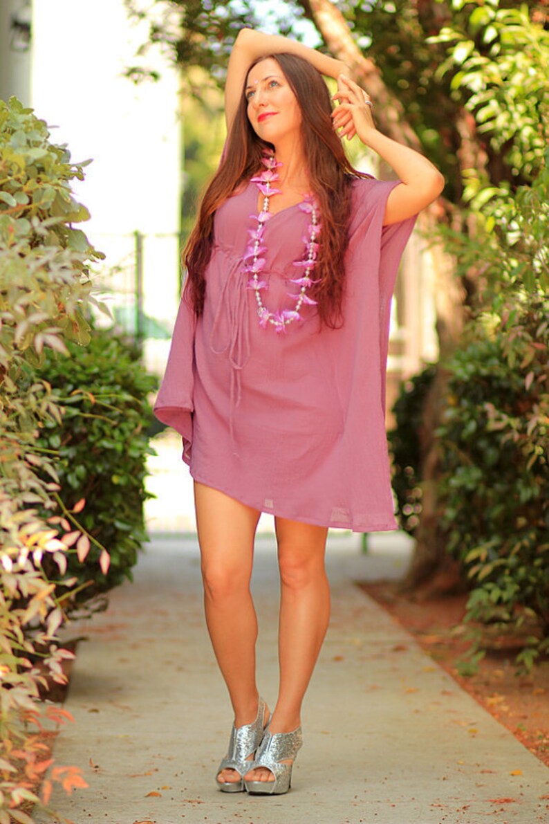May include: A woman wearing a lavender-colored dress with a pink flower garland necklace and silver glitter sandals. She is standing on a sidewalk with green bushes in the background.
