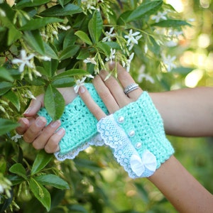 May include: A pair of mint green fingerless gloves with white lace trim and pearl buttons. The gloves are worn by a person with their hand resting on a green leafy plant.
