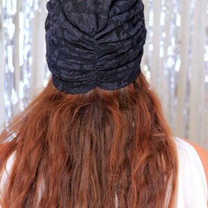 May include: A black lace turban with a gathered top. The turban is worn on a person with long, wavy, auburn hair.