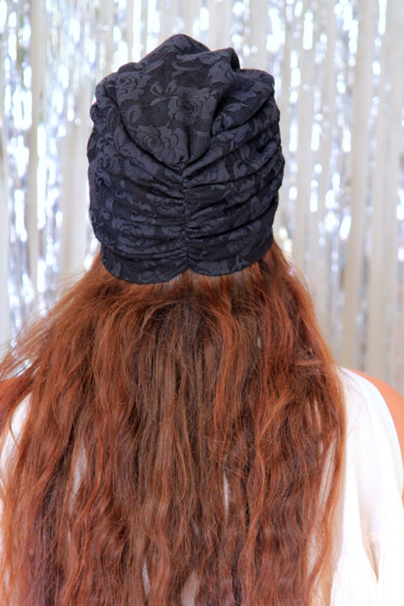 May include: A black lace turban with a gathered top. The turban is worn on a person with long, wavy, auburn hair.