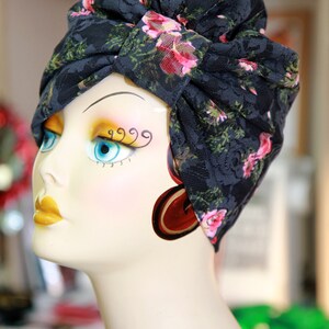 Puede incluir: Un turbante de encaje negro con estampado floral rosa. El turbante se lleva en una cabeza de maniqu&iacute; blanca con ojos azules y l&aacute;piz labial amarillo.