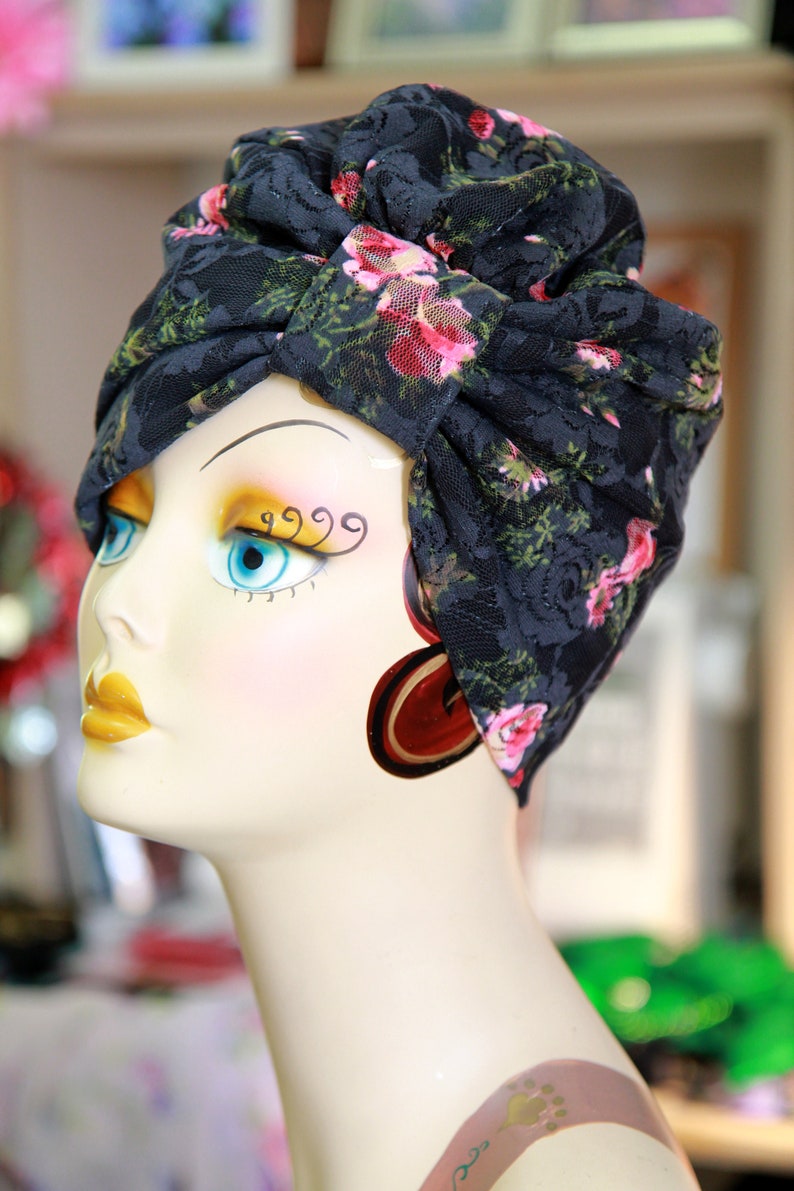 Puede incluir: Un turbante de encaje negro con estampado floral rosa. El turbante se lleva en una cabeza de maniqu&iacute; blanca con ojos azules y l&aacute;piz labial amarillo.