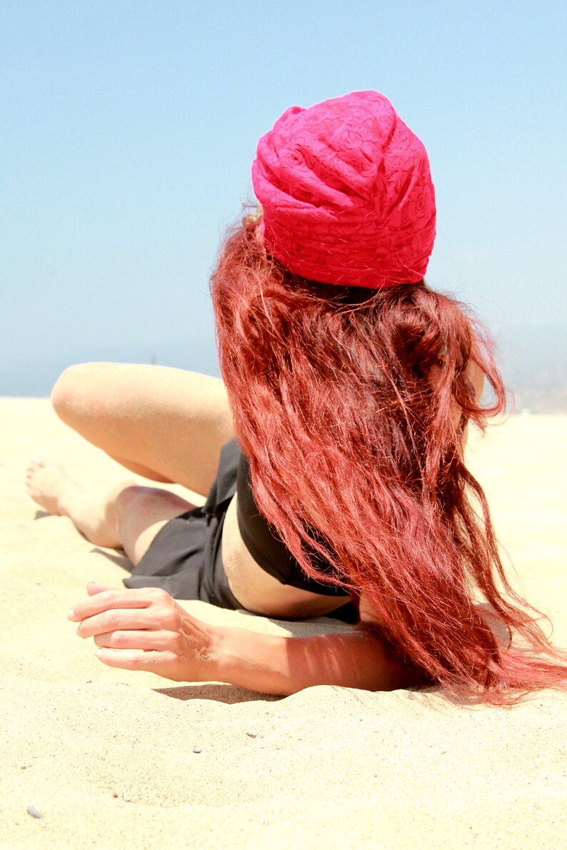 May include: A woman with long, red hair is lying on a sandy beach. She is wearing a black top and a bright pink turban.