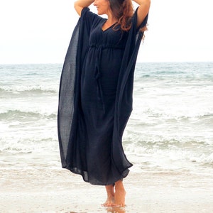 Puede incluir: Una mujer con un vestido maxi negro de silueta fluida se encuentra en una playa de arena con el oc&eacute;ano de fondo.