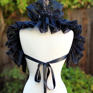 May include: A dark navy blue lace collar and shoulder piece with ruffled edges, displayed on a white mannequin. The back features a black satin ribbon tie. The background is a blurred view of green foliage.