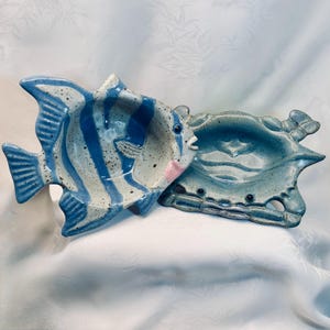 May include: Two ceramic serving dishes in the shape of sea creatures. One is a fish with blue and white stripes, and the other is a crab. The fish has a pink mouth and black eyes. The crab is a muted blue-green color.