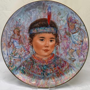 May include: Decorative plate featuring a portrait of a child with a feather in their hair, wearing a colorful beaded collar and headband. The plate has a gold rim and a background with figures in traditional attire. The artwork is detailed and colorful.