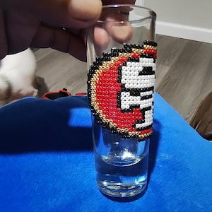 Może przedstawiać: Przezroczysty kieliszek do wódki z koralikowym wzorem. Wzór przedstawia czerwony, złoty, czarny i biały wzór. Kieliszek do wódki jest wypełniony przezroczystym płynem. Kieliszek do wódki jest trzymany przez osobę.