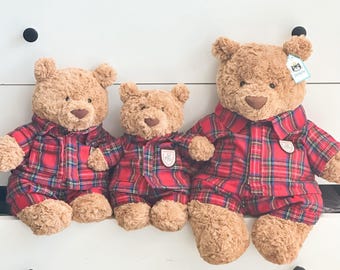 Può includere: Tre orsacchiotti di peluche marrone chiaro di diverse dimensioni, ognuno con una camicia abbottonata a quadri rossi, blu e verdi. Gli orsi hanno nasi marrone scuro e pelliccia morbida. L'orso più grande a destra ha una piccola etichetta bianca.