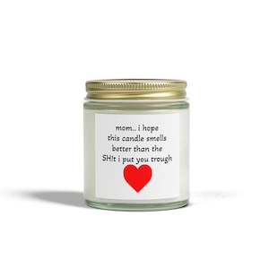 Puede incluir: Una vela en tarro de cristal con tapa dorada. La vela tiene una etiqueta blanca con el texto "mamá.. espero que esta vela huela mejor que la mierda que te hice pasar" y un corazón rojo.