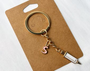 May include: A gold-coloured keyring with a syringe charm and a pink letter "S" charm. The syringe charm is white with gold accents. The keyring is attached to a brown cardboard backing.