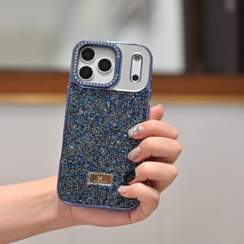 May include: A sparkling blue phone case with a glittery, textured surface. The case has a silver camera frame with a ring of clear crystals. A small, gold-colored rectangular emblem is visible on the lower part of the case.