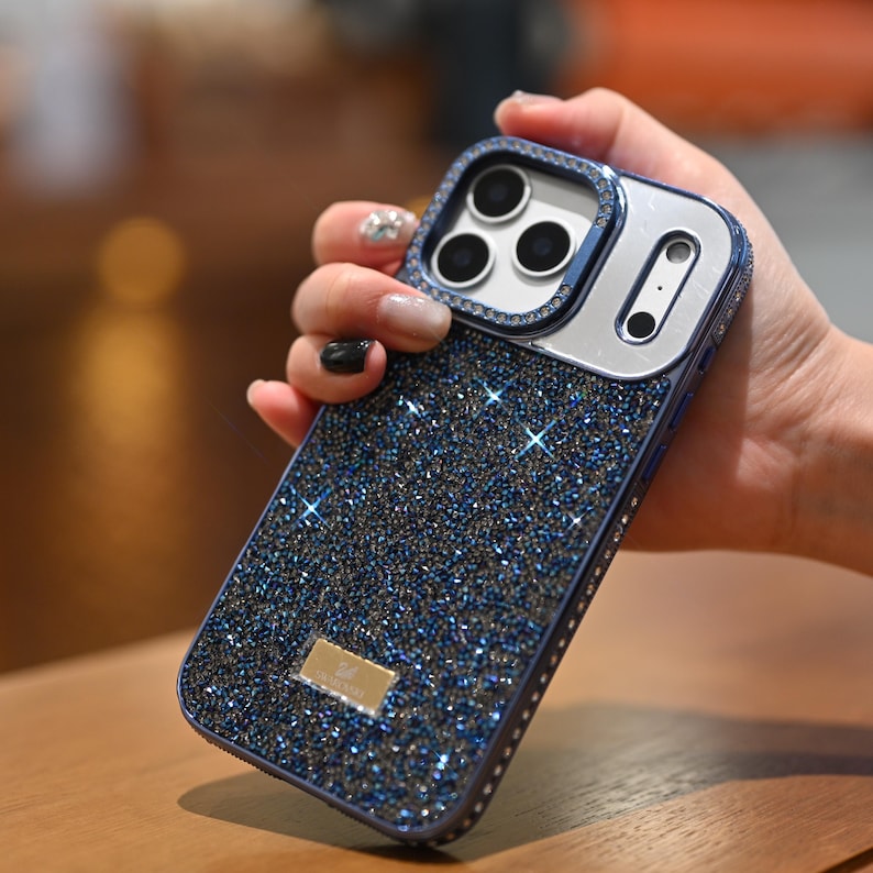 May include: A sparkling blue phone case with a glittery surface and a silver camera frame. The case has a rectangular shape and is embellished with small, shimmering crystals. A small, gold-colored logo is visible on the lower part of the case.