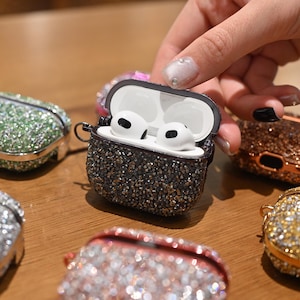 May include: A black and silver bedazzled AirPod case is open, revealing white earbuds. Several other jewel-encrusted cases in various colors, including green, pink, and gold, are scattered around. The cases are on a wooden surface.