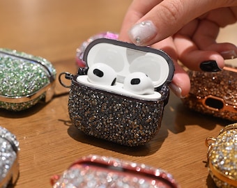 May include: A black and silver bedazzled AirPod case is open, revealing white earbuds. Several other jewel-encrusted cases in various colors, including green, pink, and gold, are scattered around. The cases are on a wooden surface.