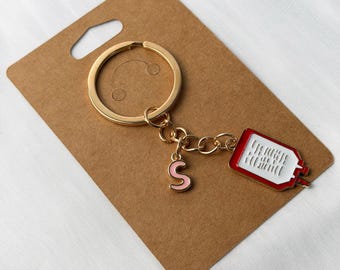 May include: Gold-coloured keyring with a red and white medical-themed charm and a pink letter "S" charm. The keyring is attached to a brown cardboard display card.