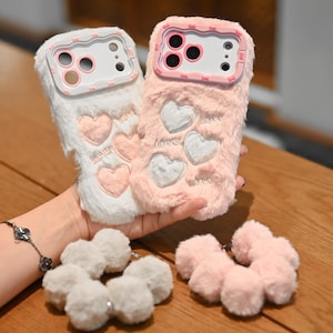 Può includere: Due custodie per telefono soffici, una bianca e una rosa, con disegni a cuore e la parola "Love" ricamata. Le custodie hanno una consistenza morbida e pelosa. Sono visibili anche due set di ciondoli a pompon abbinati.