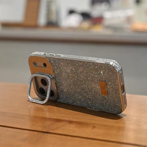 May include: A silver and glittery phone case with a ring holder. The case has a sparkly, textured back and is embellished with small, clear rhinestones around the edges and ring holder. The phone case is resting on a wooden surface.