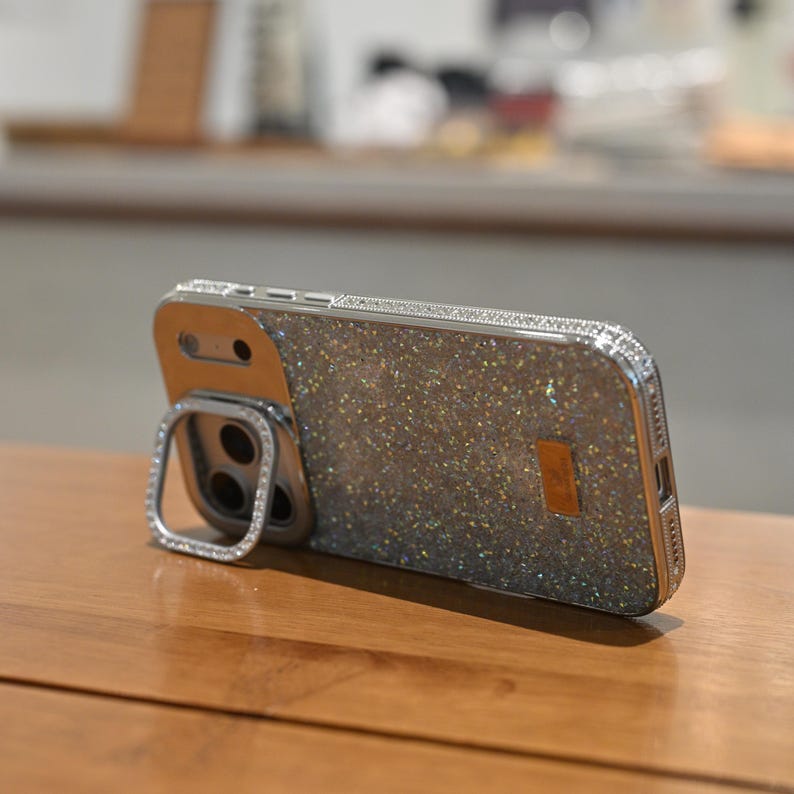 May include: A silver and glittery phone case with a ring holder. The case has a sparkly, textured back and is embellished with small, clear rhinestones around the edges and ring holder. The phone case is resting on a wooden surface.