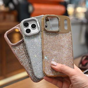 May include: Three sparkly phone cases in rose gold, silver, and gold. Each case has a glittery finish and a decorative frame around the camera lenses. The cases are held in a hand, showcasing their shimmering appearance.
