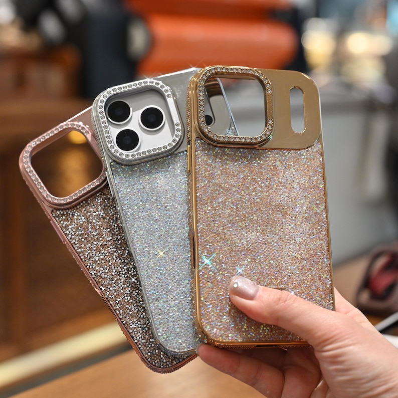 May include: Three sparkly phone cases in rose gold, silver, and gold. Each case has a glittery finish and a decorative frame around the camera lenses. The cases are held in a hand, showcasing their shimmering appearance.
